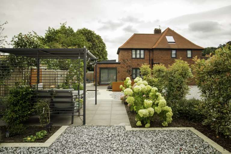 A landscaped garden with gravel paths, patio seating under a pergola, flowering shrubs, and a brick house with a red roof in the background showcases thoughtful architectural design.