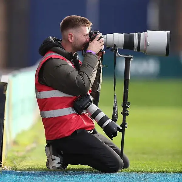 Nathan Stirk, professional photographer, kneeling beside a sports field with a telephoto camera